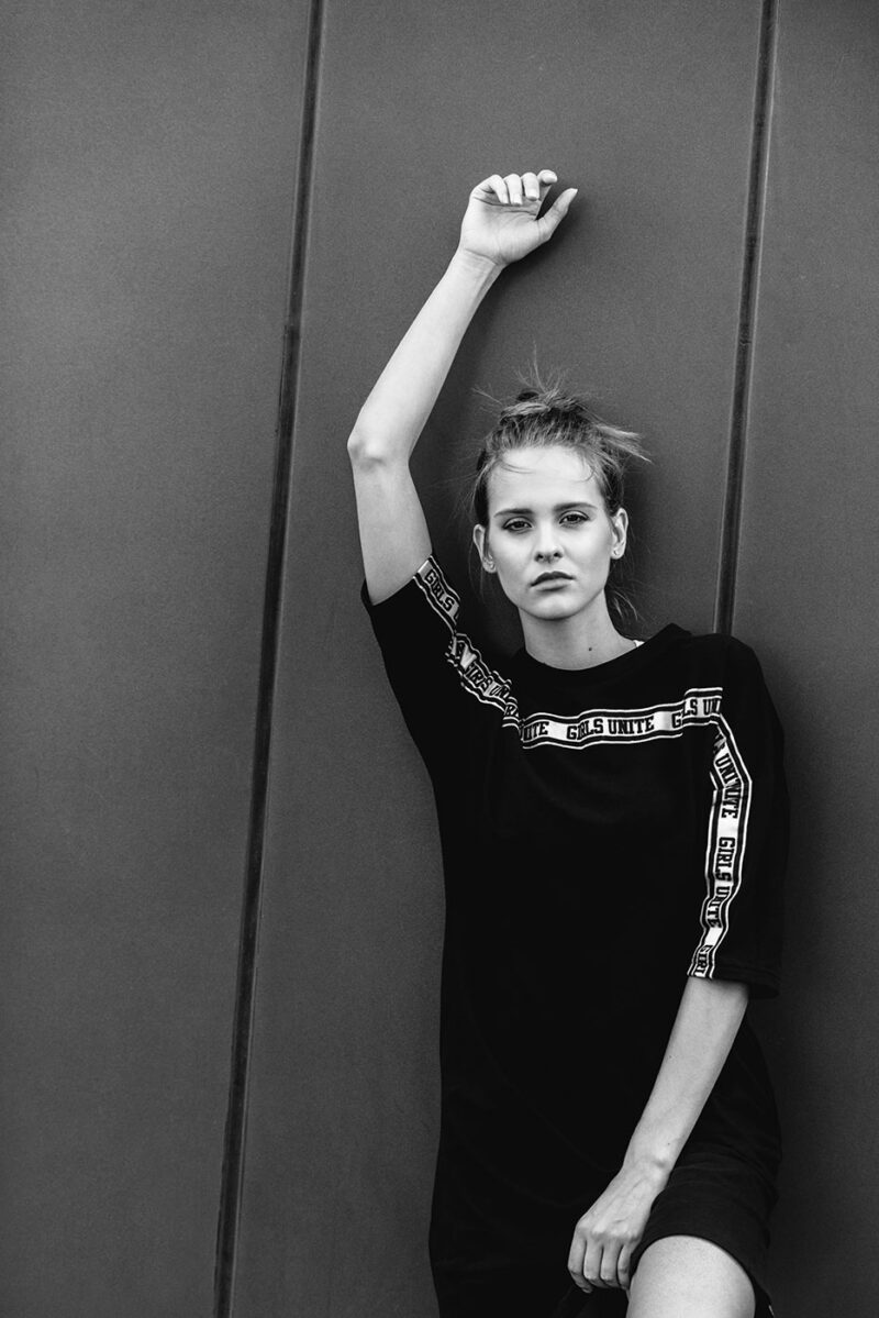 Девушка позирует в темном платье-футболке, стоя у тёмной стены и высоко подняв руку