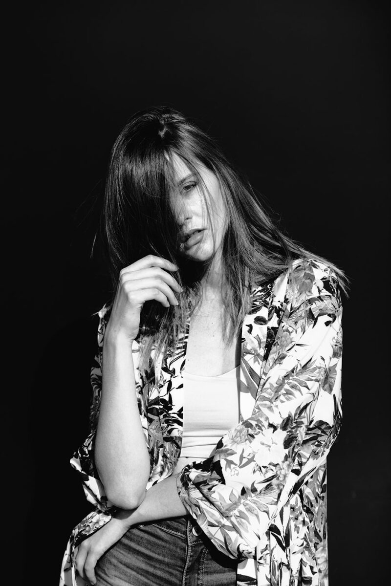 Чёрно-белый поясной портрет девушки в накидке с цветочным принтом, прикрывающей лицо волосами на черном фоне