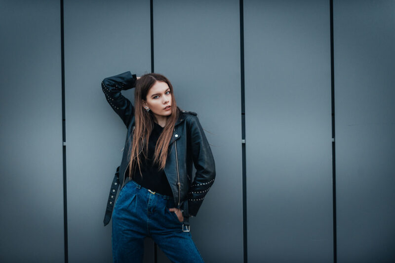Девушка в кожаной куртке стоит у серой стены современной постройки, касаясь рукой головы