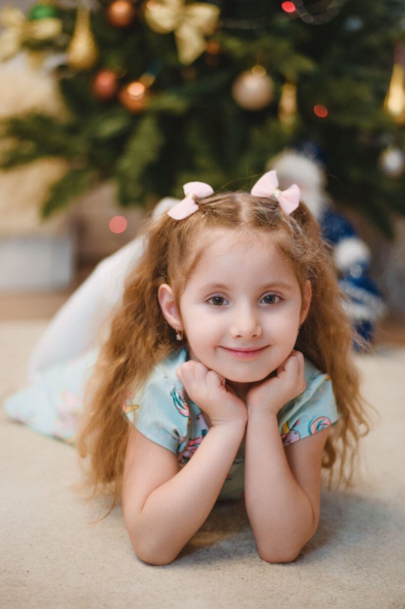 Девочка лежит на животе и улыбается перед новогодней ёлкой, домашняя фотосессия