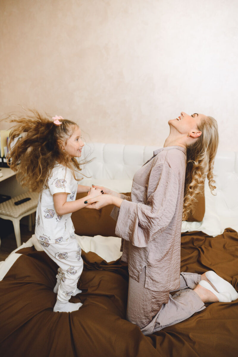 Девочка прыгает на кровати, держась за руки с мамой
