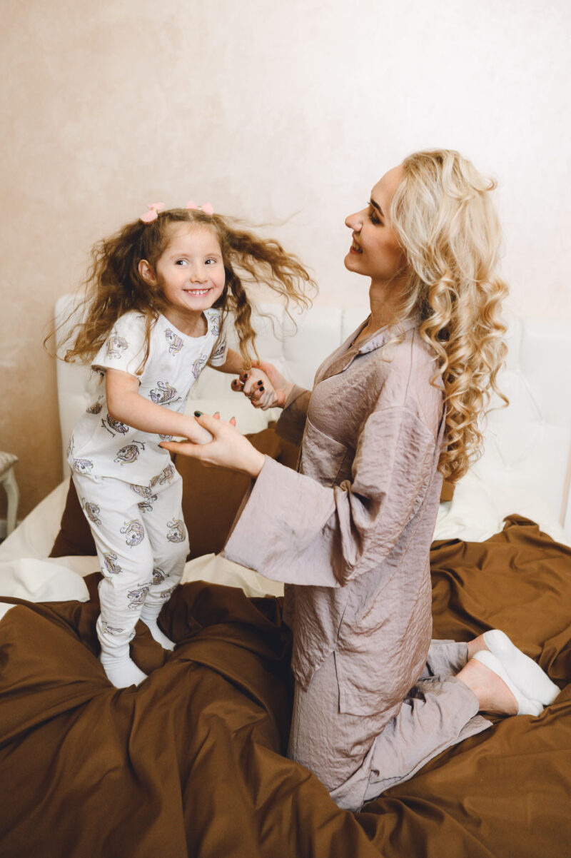 Дочка подпрыгивает на кровати, мама держит её за руки в спальне