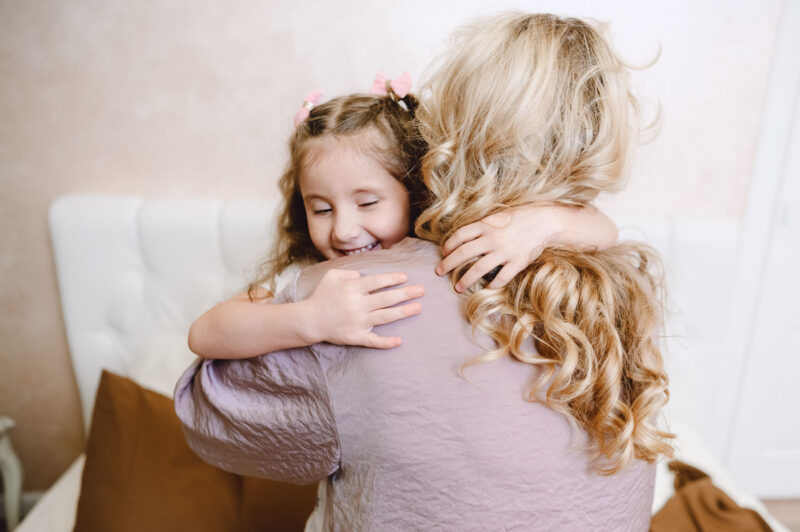 В уютной квартире девочка обнимает маму за шею, вид со спины