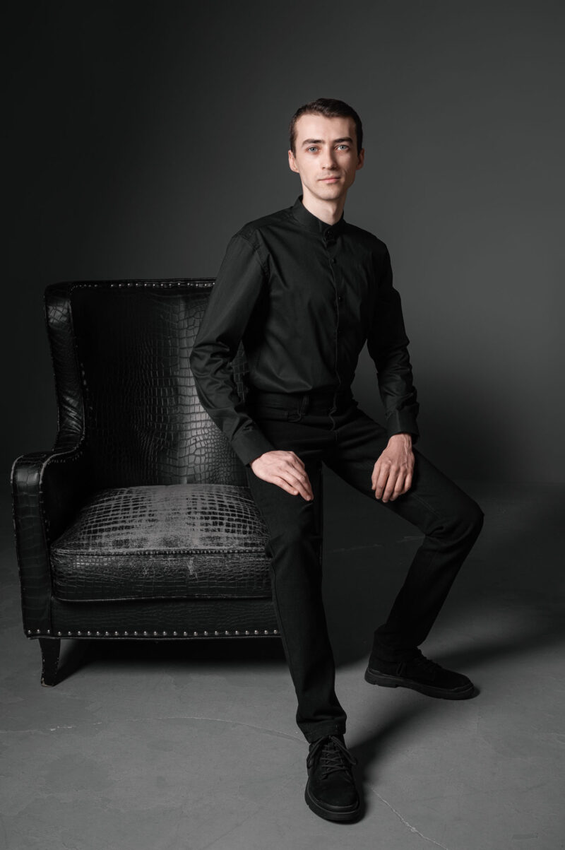 Мужчина в чёрной рубашке сидит на подлокотнике кресла на сером фоне