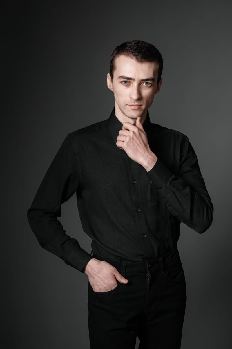 Мужской портрет в чёрной рубашке с рукой у подбородка на тёмном фоне