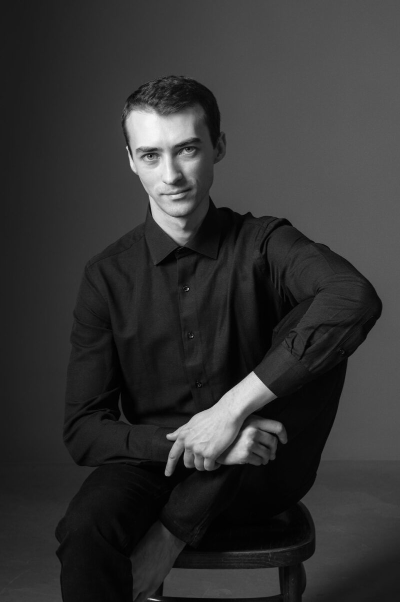 Мужчина в чёрной рубашке сидит на стуле, взгляд в камеру, чёрно-белый кадр