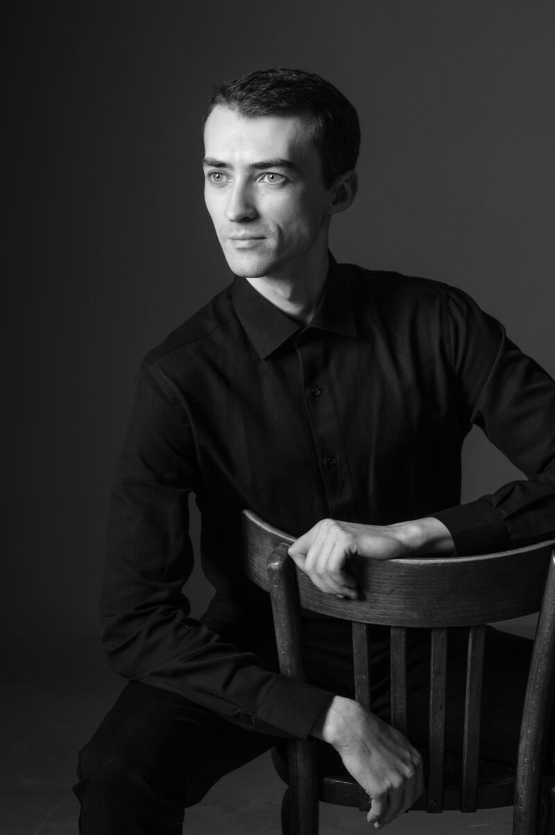 Мужчина сидит на деревянном стуле на тёмном студийном фоне, смотря в сторону