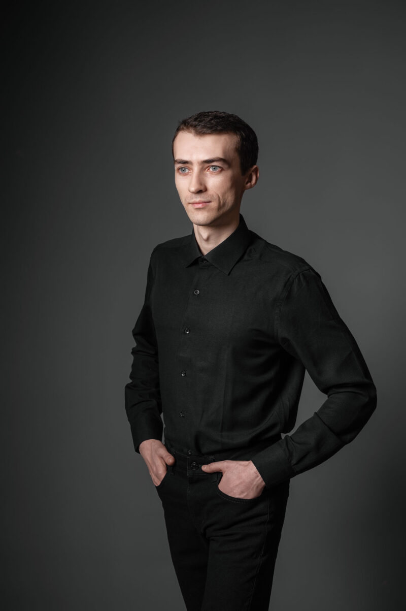 Мужчина стоит в тёмном зале фотостудии, руки в карманах брюк