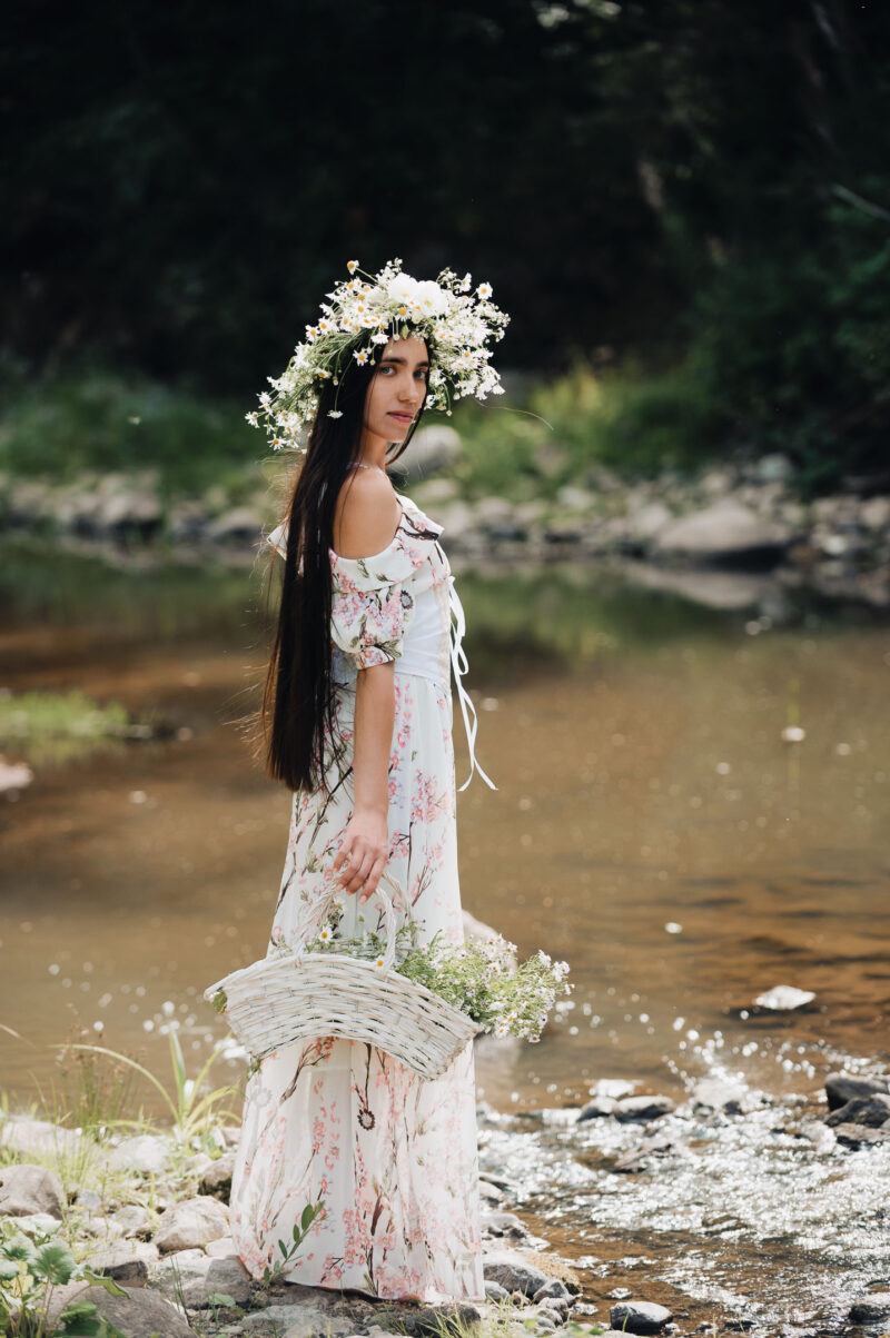 портрет девушки с венком на голове и с плетёной корзинкой в лёгком летнем платье на природе на фоне лесной реки