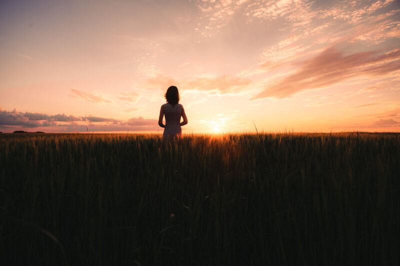 Силуэт девушки посреди бескрайнего поля под оранжевым небом заката
