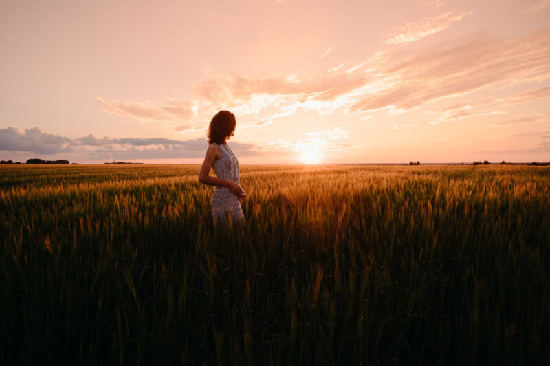 Женский портрет в полный рост в лучах контрового света на фоне заходящего солнца на поле