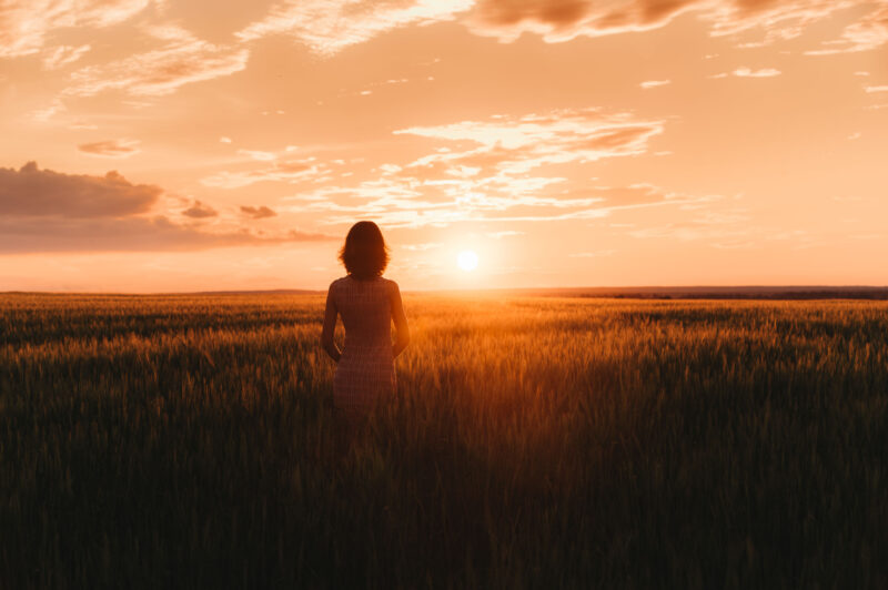 На фоне горизонта девушка стоит спиной к камере, наблюдая за закатом в поле под Ярославлем