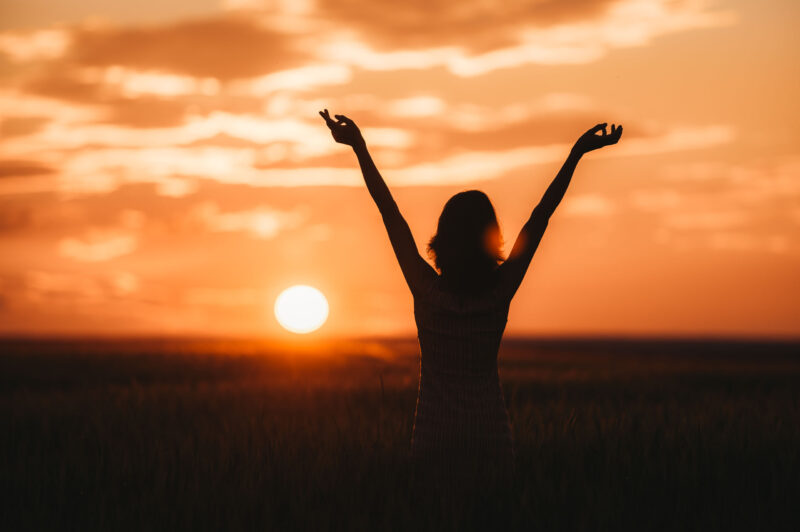 Поднявшая руки вверх девушка позирует на фоне заходящего солнца в открытом поле