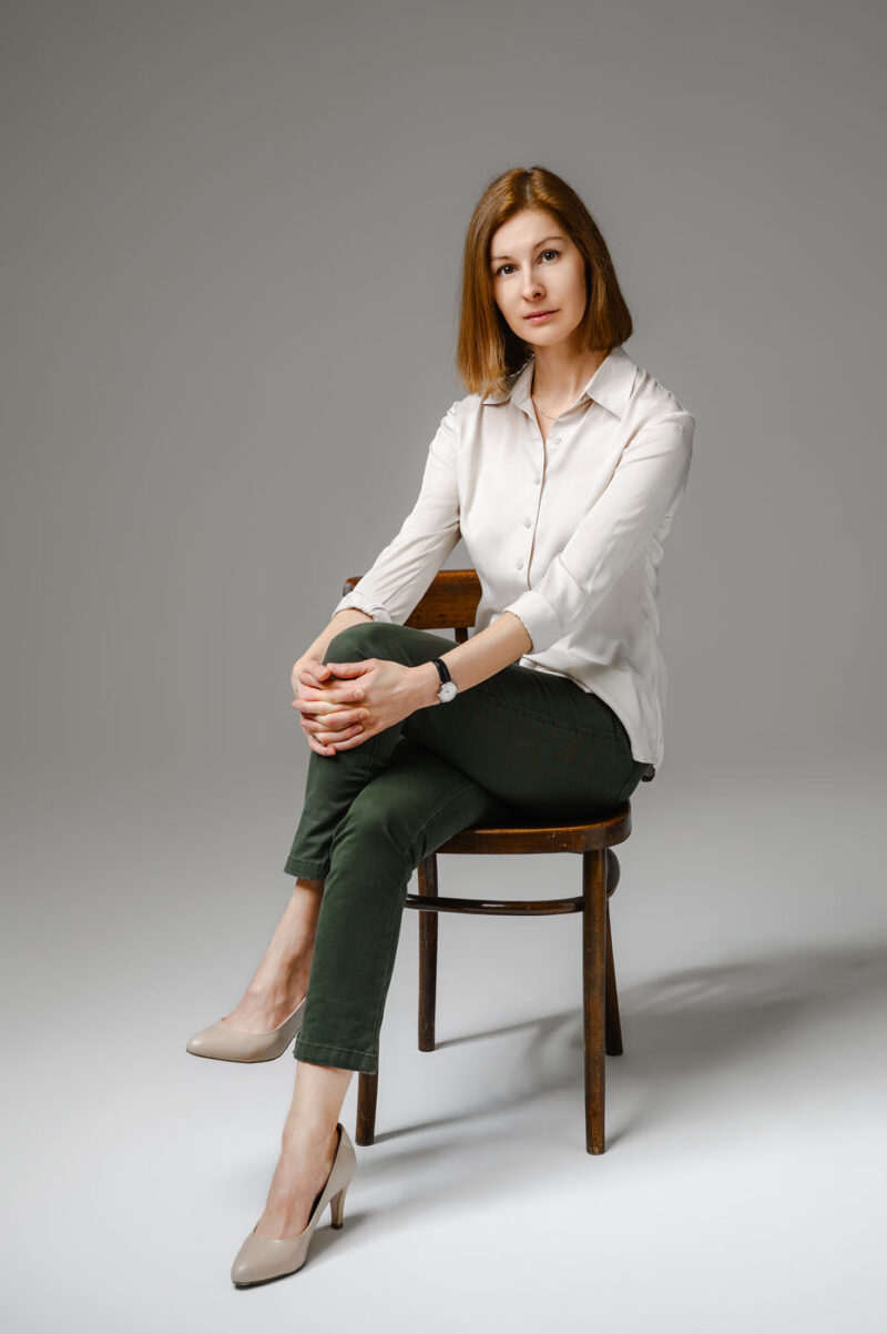 Сидящая на деревянном стуле девушка в светлой блузке и туфлях на фоне студийной циклорамы