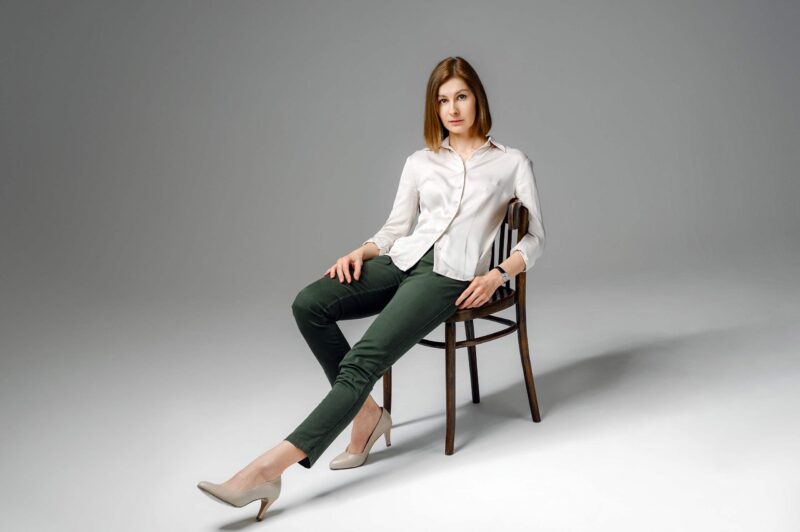 На фоне серой стены циклорамы девушка сидит на стуле, вытянув одну ногу вперед