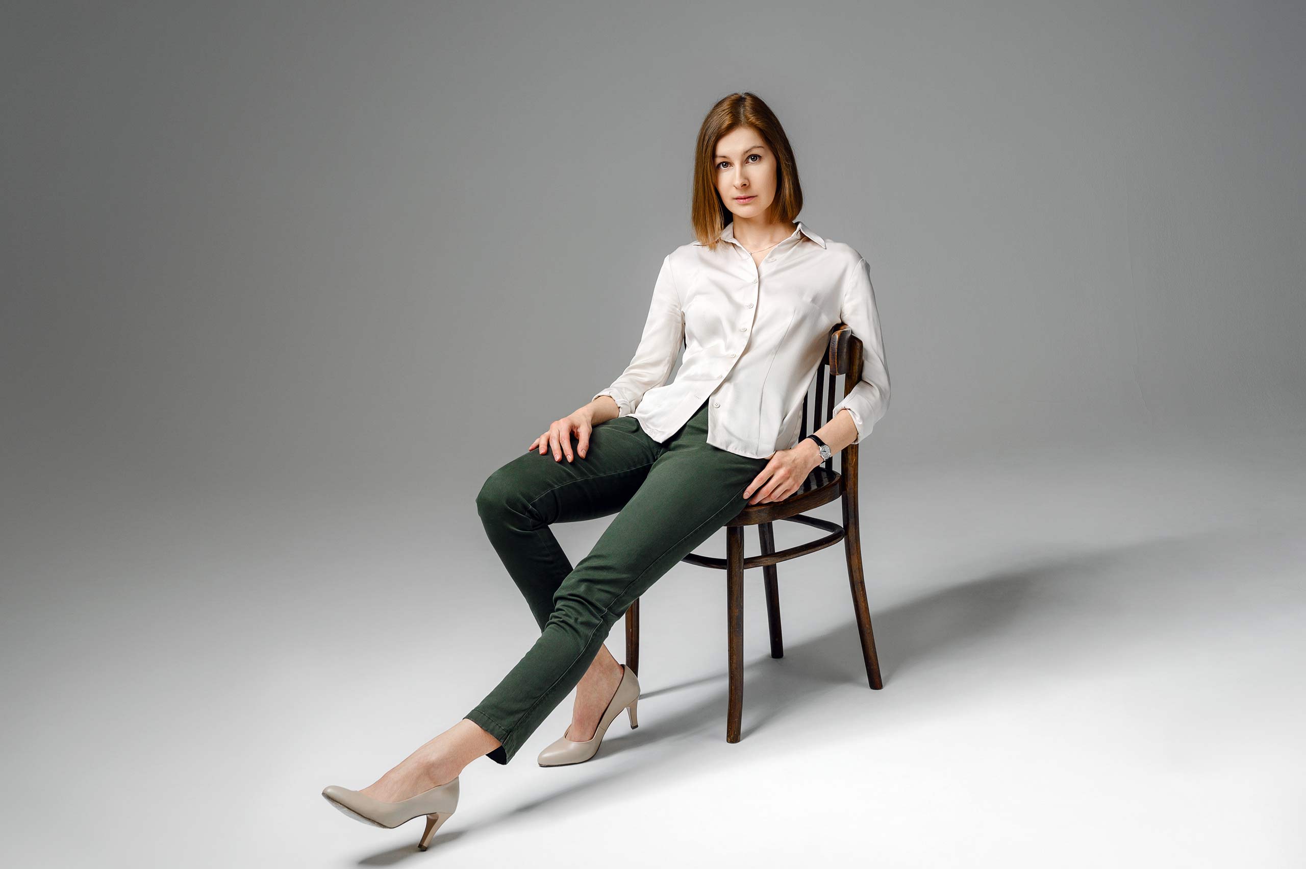 На фоне серой стены циклорамы девушка сидит на стуле, вытянув одну ногу вперед