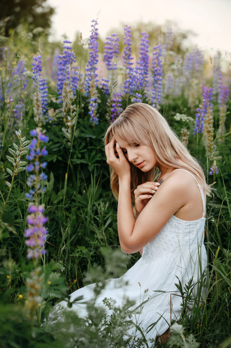 Девушка в белом платье сидит среди цветущих люпинов при естественном дневном свете