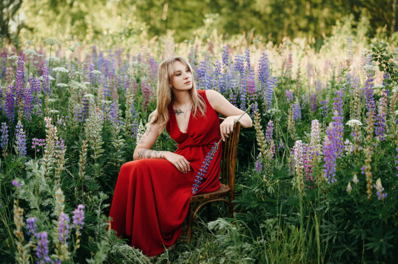 Девушка в красном платье сидит на стуле среди люпинов в мягком свете заката