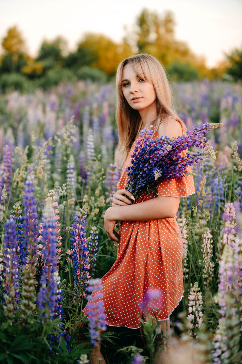 Девушка в оранжевом платье с букетом на фоне закатного люпинового поля