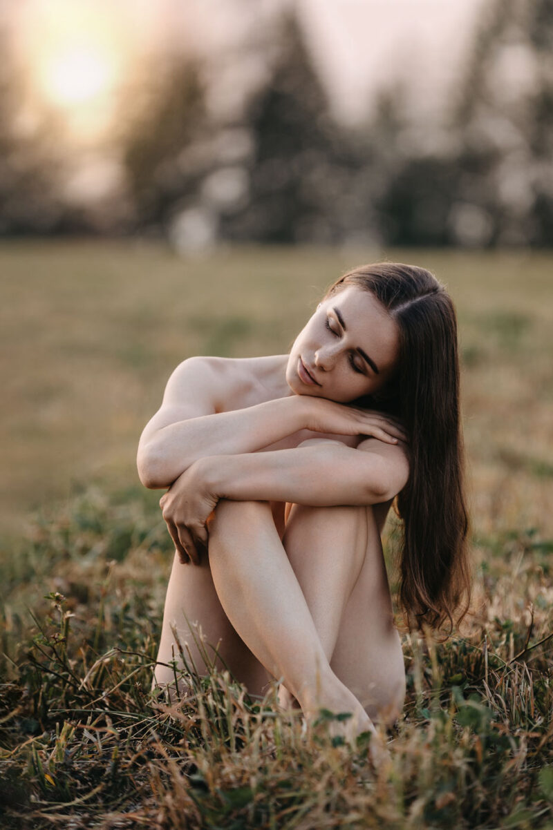 Обнажённая девушка сидит в поле, освещённая мягким светом заходящего солнца